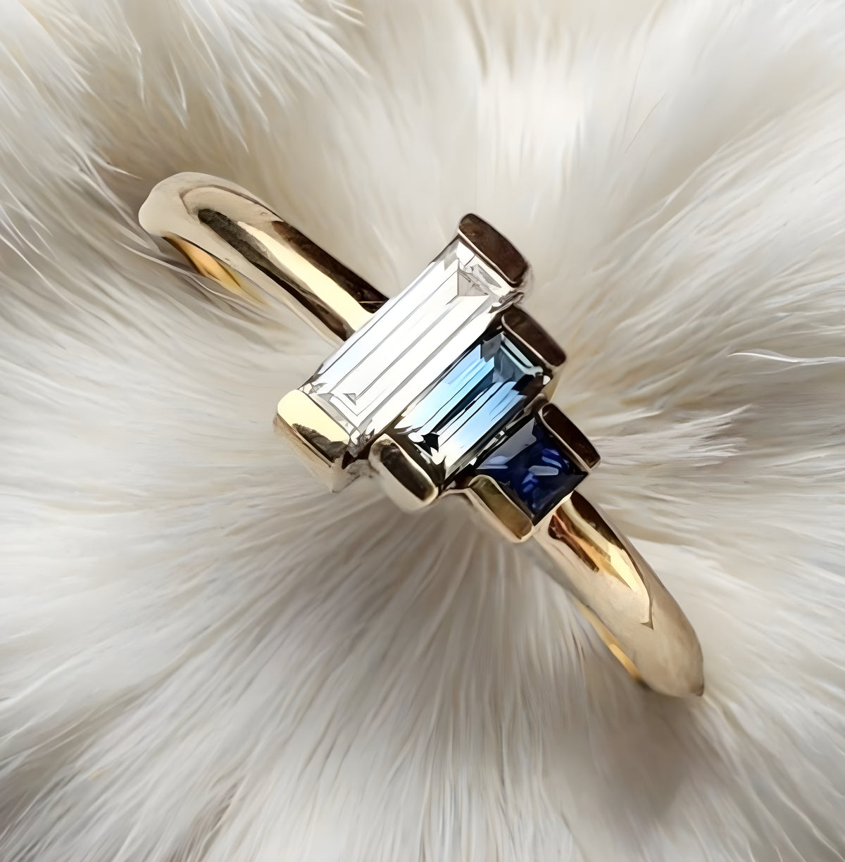 Gold ring with emerald-cut and blue gemstones on a white fur background